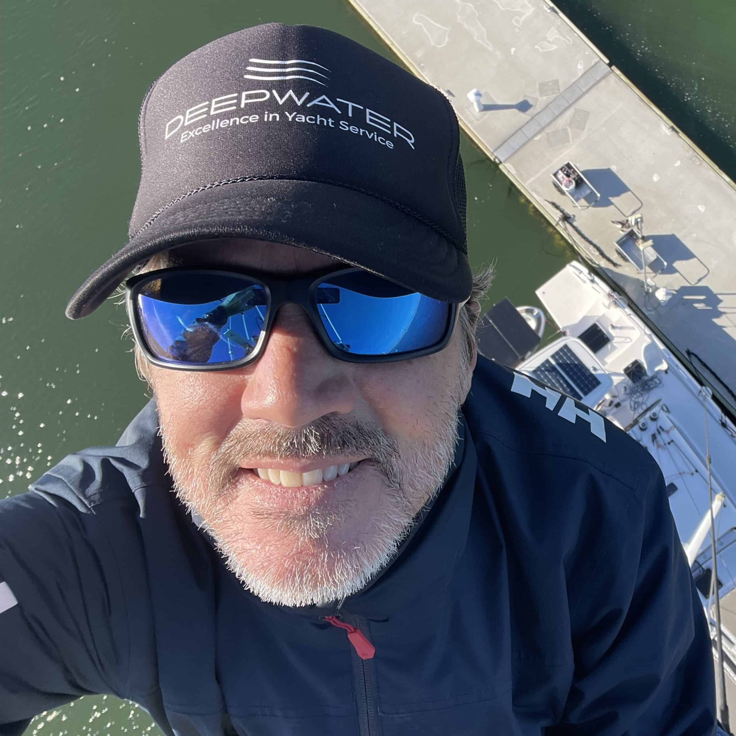 Man wearing sunglasses and a "Deepwater" cap smiles at the camera while positioned high above a yacht docked at a marina.