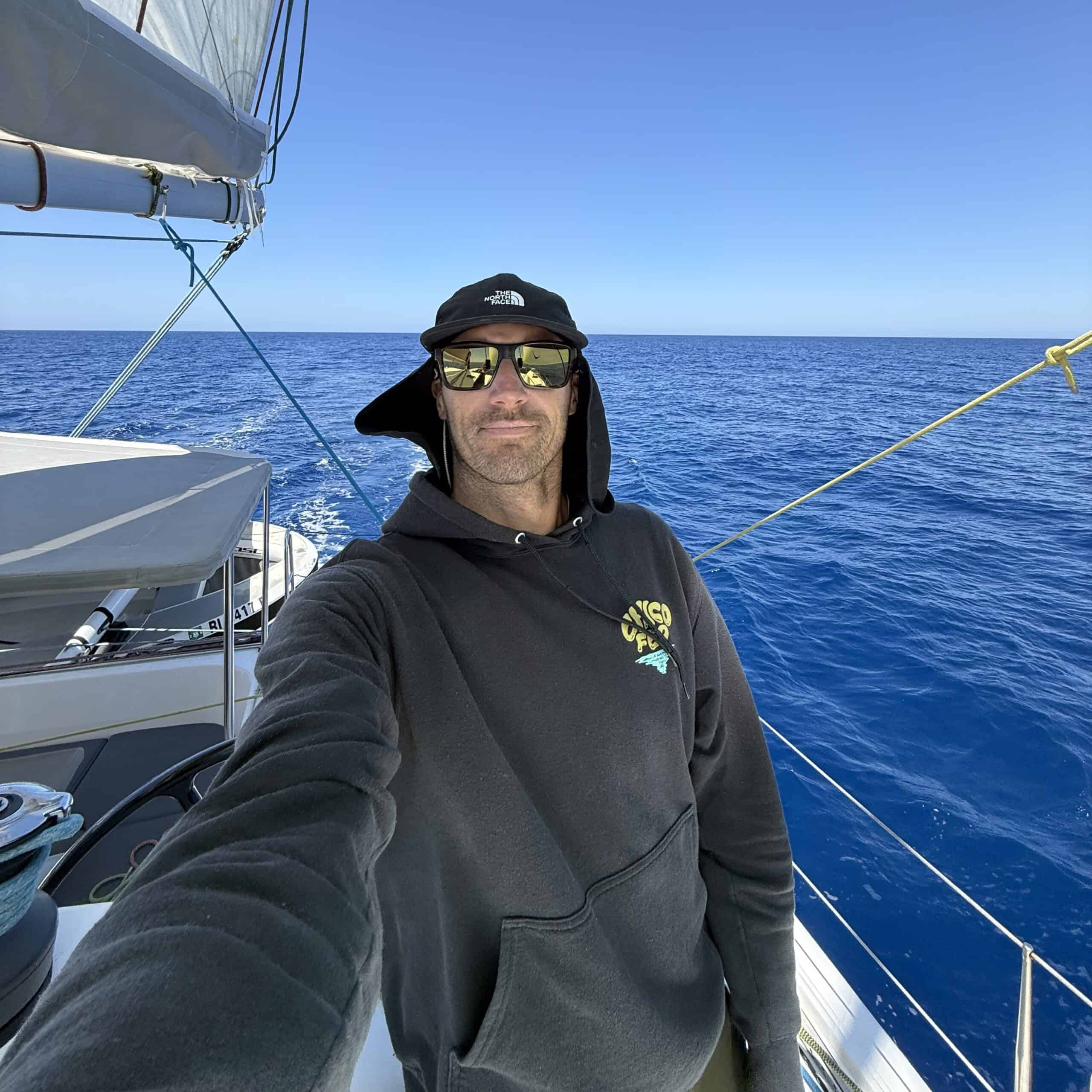 A person wearing sunglasses and a black hoodie stands on a sailboat in the ocean, with clear blue water and sky in the background.