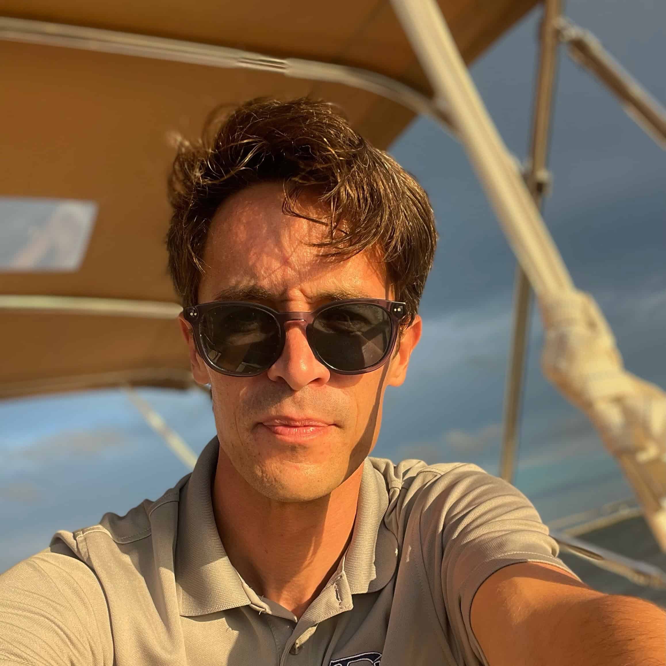 A man wearing sunglasses and a gray polo shirt sits on a boat under a canopy, with sunlight illuminating his face.