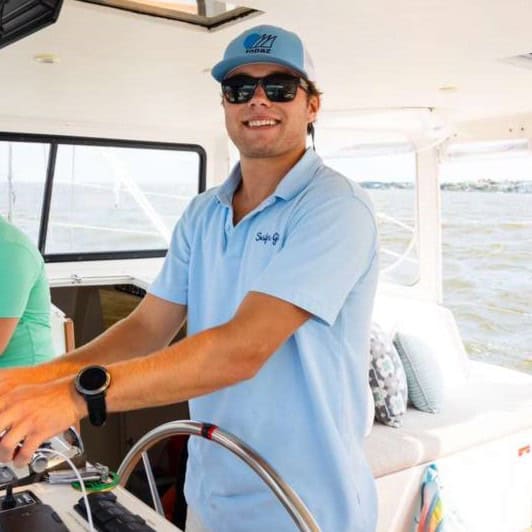 A man wearing sunglasses and a blue polo shirt steers a boat inside a cabin, smiling at the camera.